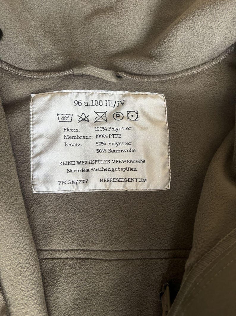 May include: A white label with black text and symbols. The label reads "96 u.100 III/IV", "40&deg;", "Fleece: 100% Polyester", "Membrane: 100% PTFE", "Besatz: 50% Polyester 50% Baumwolle", "KEINE WEICHSP&Uuml;LER VERWENDEN!", "Nach dem Waschen gut sp&uuml;len", "FECSA/2017", "HEERESEIGENTUM".