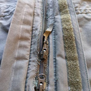 May include: Close-up of a worn, olive-green jacket zipper. The metal zipper is partially unzipped, revealing the teeth. The fabric around the zipper shows signs of wear and tear, with frayed edges and a faded appearance. A strip of hook-and-loop fastener is visible.