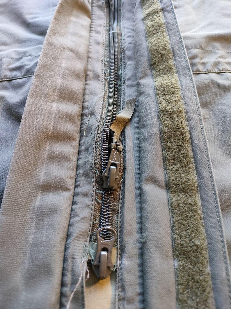 May include: Close-up of a worn, olive-green jacket zipper. The metal zipper is partially unzipped, revealing the teeth. The fabric around the zipper shows signs of wear and tear, with frayed edges and a faded appearance. A strip of hook-and-loop fastener is visible.