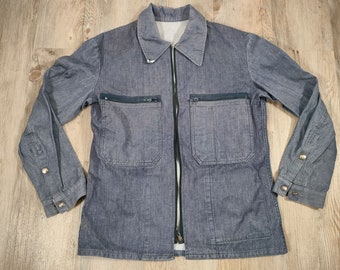 May include: A gray denim jacket with a zipper closure and two large pockets on the front. The jacket has a buttoned cuff on each sleeve.