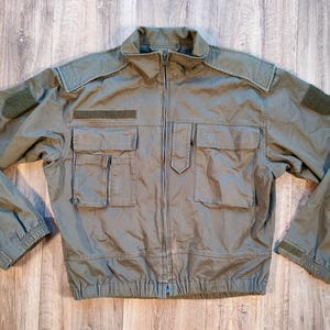 May include: Olive green military-style jacket with a full-length zipper. The jacket features multiple pockets, including two chest pockets and two lower pockets. It has a stand-up collar, shoulder epaulets, and Velcro patches on the sleeves. The jacket is made of a durable, possibly water-resistant material.