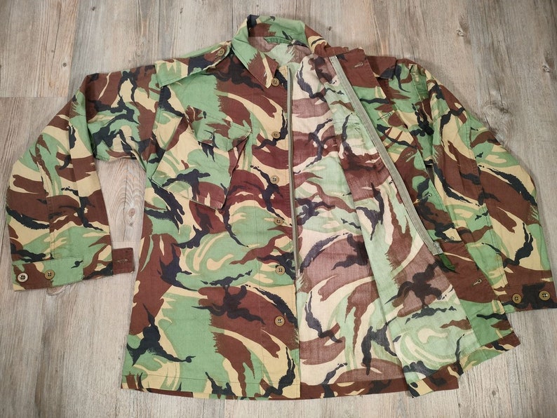 May include: A camouflage-patterned shirt with a zip closure and button-up front. The shirt has two large pockets on the front and a green zip pull.