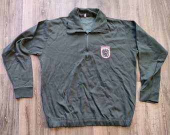 Può includere: Una felpa a maniche lunghe verde scuro con chiusura a zip e una piccola toppa ricamata sul petto. La toppa presenta uno stemma con il testo "Bundeswehr" in bianco.