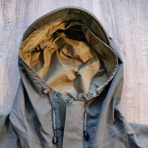 May include: Close-up of a green hooded jacket. The hood is open, revealing a lighter green interior. The jacket has a drawstring and a zipper closure. The fabric appears to be a durable, weather-resistant material, suitable for outdoor wear.