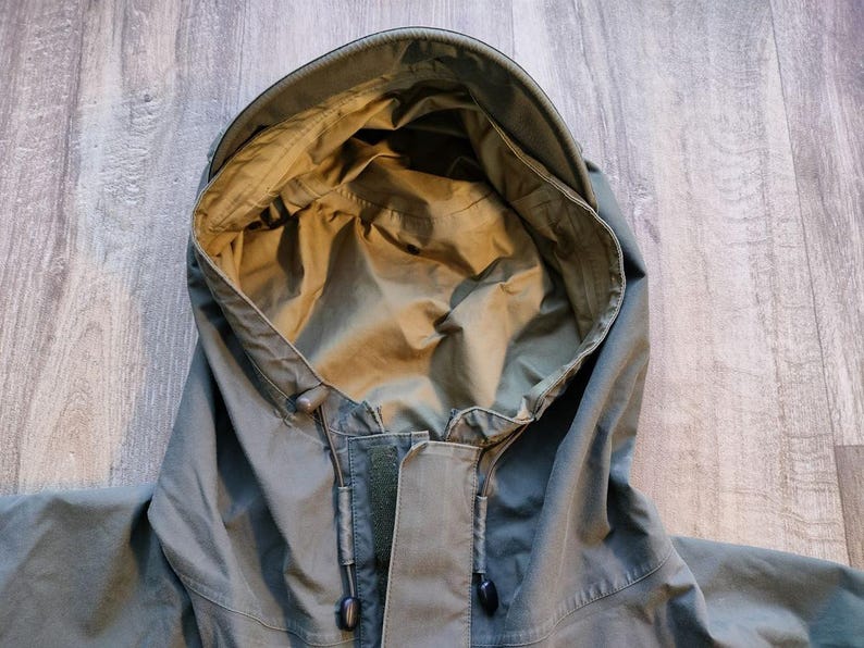 May include: Close-up of a green hooded jacket. The hood is open, revealing a lighter green interior. The jacket has a drawstring and a zipper closure. The fabric appears to be a durable, weather-resistant material, suitable for outdoor wear.