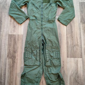 Puede incluir: Mono verde de estilo militar con múltiples bolsillos, cremalleras y cierre de cremallera frontal. La prenda de manga larga tiene cuello y un parche en el pecho. El mono está tendido sobre una superficie de madera.