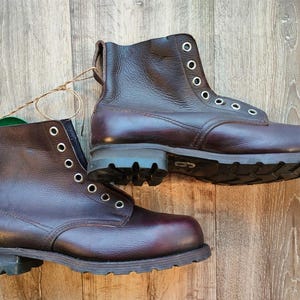 Op de afbeelding: Een paar bordeauxrode leren werklaarzen met metalen vetergaten en zolen met noppen. De laarzen zijn nieuw, met een aangehecht label. De laarzen staan op een houten ondergrond.
