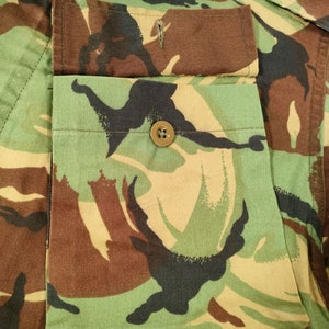May include: Close-up of a camouflage fabric with a flap pocket and a button. The pocket is sewn onto the fabric and has a brown button.