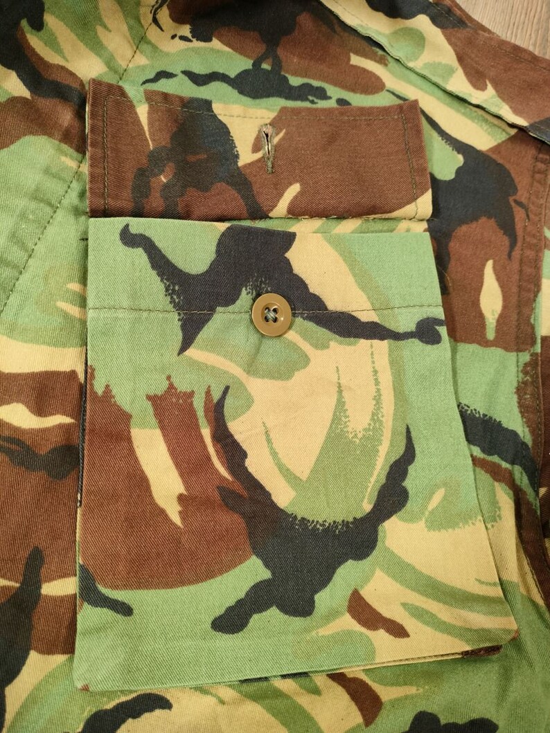 May include: Close-up of a camouflage fabric with a flap pocket and a button. The pocket is sewn onto the fabric and has a brown button.