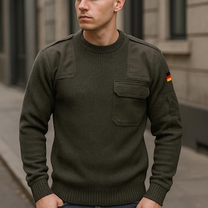 Może przedstawiać: Zielony sweter z łatą na ramieniu i kieszenią z przodu. Sweter ma żebrowaną fakturę i okrągły dekolt. Łata ma małą flagę niemiecką.