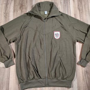 May include: A green zippered jacket with a German eagle logo on the left chest. The jacket has a high collar and long sleeves.