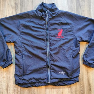 Peut inclure: Veste zippée bleu marine avec un logo de chat rouge et du texte. La veste a un col montant, des poignets élastiques et une fermeture éclair intégrale. Le texte sur la veste indique "XV" et "So Big Feet". La marque "motif" est également visible.