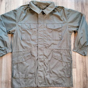 Op de afbeelding: Olijfgroene jas in militaire stijl met een kraag en meerdere voorzakken. De jas heeft lange mouwen en een taille met trekkoord. De stof lijkt een licht, duurzaam materiaal te zijn. De jas ligt plat op een houten oppervlak.