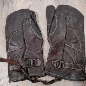 May include: A pair of brown leather mittens with a strap and buckle closure. The mittens have a white stitching detail.