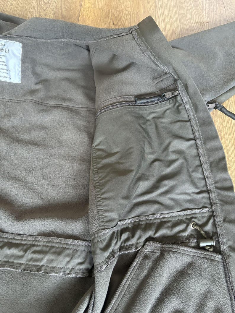 May include: A dark green fleece jacket with a zipper closure and multiple pockets. The jacket has a fleece lining and a drawstring at the waist.