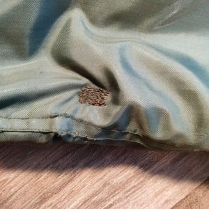 以下が含まれることがあります： 緑と青の色合いの生地のクローズアップ。生地はわずかな光沢があり、テクスチャが見えます。小さな茶色の不規則なパッチが生地に見え、修復または損傷を示しています。生地の端が見えます。