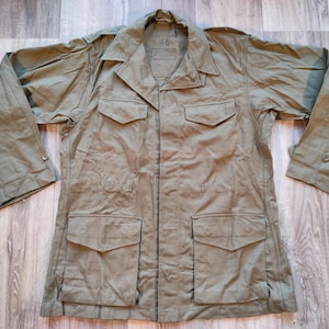 May include: A green utility jacket with a button-up front, two large pockets on the front, and two smaller pockets on the chest. The jacket has a collar and long sleeves.