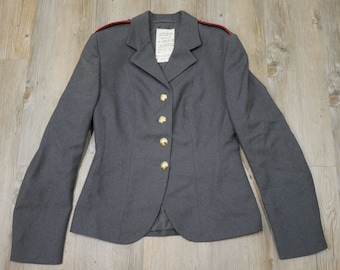 Könnte beinhalten: Ein grauer Blazer mit roten Paspeln an den Schultern und goldenen Knöpfen. Der Blazer hat einen klassischen Stil und ist aus hochwertigem Stoff gefertigt.