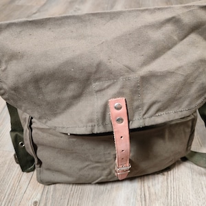 Puede incluir: Un bolso de mensajero de lona verde con una correa de cuero marrón y una hebilla. El bolso tiene un cierre de solapa y un pequeño bolsillo en la parte delantera.