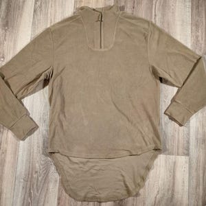 Könnte beinhalten: Ein langärmliges, beiges Hemd mit hohem Kragen und einem Reißverschluss vorne. Das Hemd hat einen langen, abgerundeten Saum.