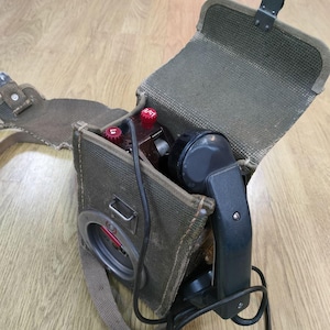 May include: A vintage military field telephone with a black handset and a green canvas carrying case. The phone has a red dial and a black cord.
