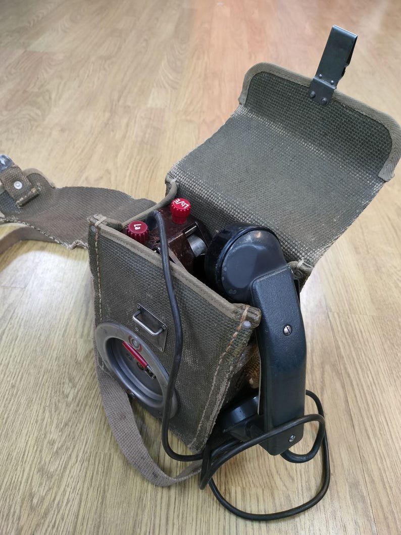 May include: A vintage military field telephone with a black handset and a green canvas carrying case. The phone has a red dial and a black cord.