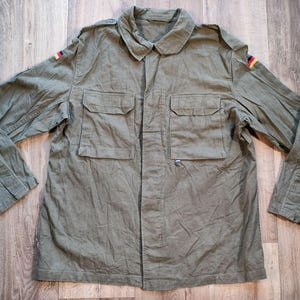 May include: Olive green military-style jacket with four front pockets and a button closure. The jacket features the German flag on both shoulders. The long-sleeved jacket is made of a durable fabric and has a collared neckline.