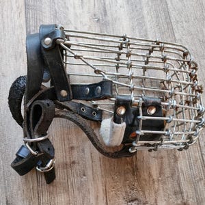 May include: A dog muzzle featuring a metal wire cage and black leather straps. The cage is rectangular, and the leather straps have buckles and rivets for adjustment. A small, light-coloured pad is attached to the muzzle.