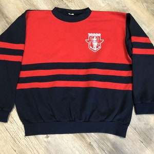 Pode incluir: Uma camisa de frio vermelha e azul marinho com um logotipo bordado branco na frente. O logotipo apresenta um escudo com um paraquedas e uma âncora dentro. A camisa de frio tem listras vermelhas nas mangas e no peito.