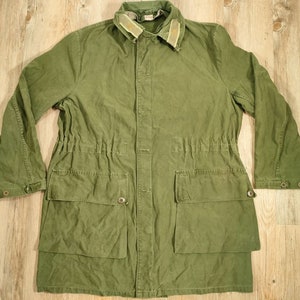 Peut inclure: Une veste en toile verte avec une taille à cordon et plusieurs poches. La veste a un col en velours côtelé marron.