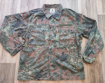 Puede incluir: Una camisa de manga larga de camuflaje verde, marrón y negro con cierre de botones en la parte delantera y dos bolsillos de solapa. La camisa tiene un pequeño parche de la bandera alemana en la manga izquierda.