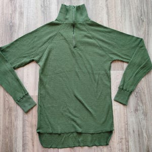 Könnte beinhalten: Olivgrünes Langarmshirt mit Rippstruktur und Stehkragen. Das Shirt hat einen Viertel-Reißverschluss am Ausschnitt und gerippte Bündchen. Der Saum ist leicht gebogen. Ein lässiges Oberteil für den Alltag.