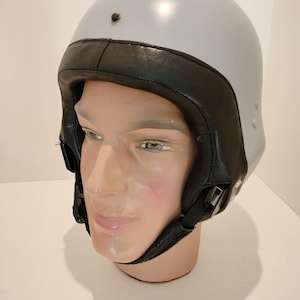 Peut inclure: Un casque de moto gris avec une doublure en cuir noir et une sangle de menton. Le casque a une finition lisse et brillante et un petit bouton noir sur le dessus.