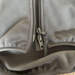 May include: Close-up of a dark gray fleece jacket with a zipper closure. The zipper is partially open, revealing the fleece lining. The jacket has a pocket on the left side.