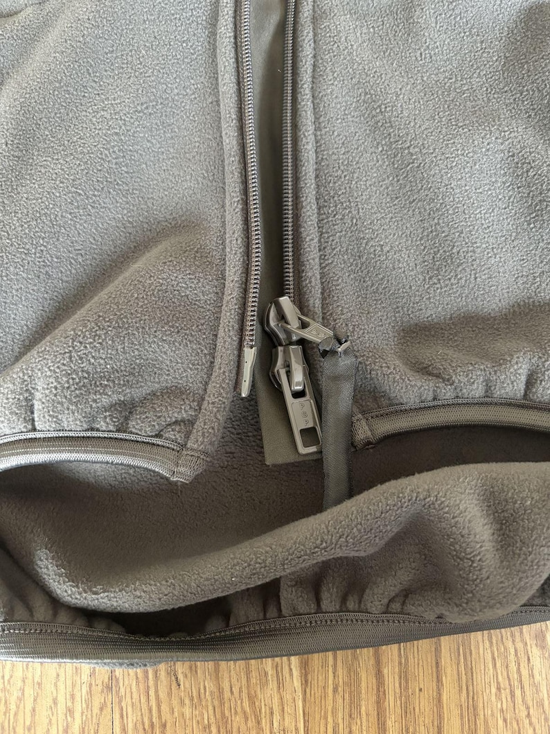 May include: Close-up of a dark gray fleece jacket with a zipper closure. The zipper is partially open, revealing the fleece lining. The jacket has a pocket on the left side.
