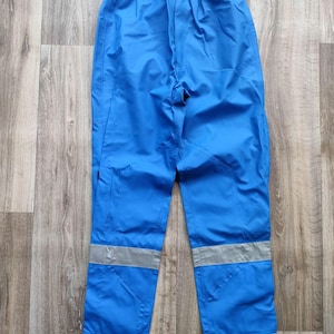 May include: Bright blue waterproof pants with reflective silver stripes near the ankles. The pants have an elastic waistband and a drawstring. The material appears to be a lightweight, durable fabric, suitable for outdoor activities.