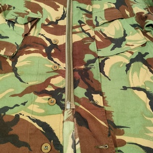 May include: A camouflage jacket with a zip fastening and multiple buttons. The jacket is made of a cotton fabric and has a green, brown, and black colour scheme.