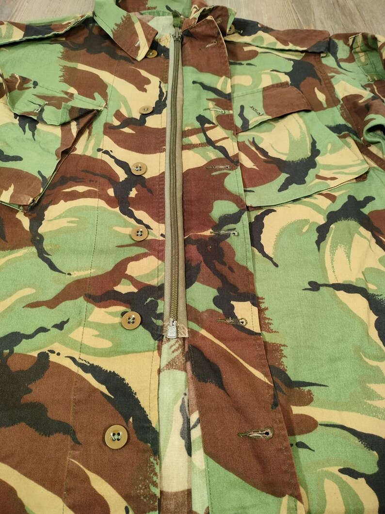 May include: A camouflage jacket with a zip fastening and multiple buttons. The jacket is made of a cotton fabric and has a green, brown, and black colour scheme.