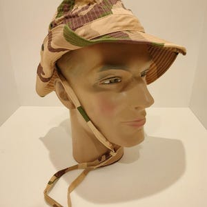 Peut inclure: Chapeau de pêcheur camouflage avec un large bord et une mentonnière. Le chapeau présente un motif beige, marron et vert. Le chapeau est présenté sur une tête de mannequin.
