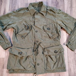 Può includere: Giacca verde oliva in stile militare con più tasche e chiusure con bottoni. La giacca ha un collo, maniche lunghe e una vita con coulisse. L'indumento è realizzato in tessuto resistente, adatto per attività all'aperto.