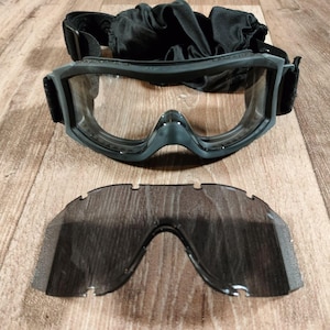 May include: Black tactical goggles with clear lenses and a spare tinted lens. The goggles have a black frame and adjustable strap. A black cloth bag is included. The items are displayed on a wooden surface.