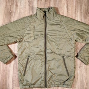 Könnte beinhalten: Olivgrüne isolierte Jacke mit durchgehendem Reißverschluss und zwei Reißverschlusstaschen. Die Jacke hat einen hohen Kragen und besteht aus einem glatten, gesteppten Material. Die Jacke liegt flach auf einer Holzoberfläche.