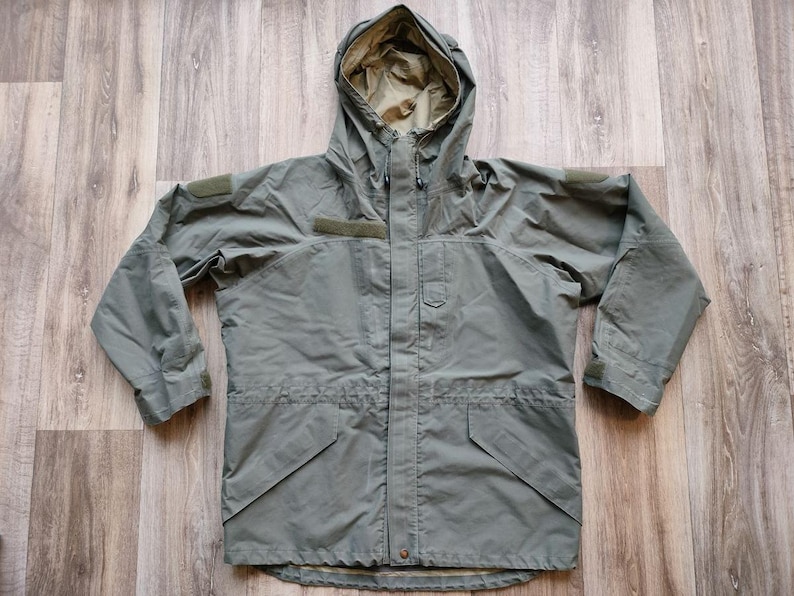 May include: A sage green hooded jacket with a drawstring waist and multiple pockets. The jacket has a full-length zipper and patches on the sleeves. The hood is lined with a lighter green fabric. This outerwear is suitable for outdoor activities.