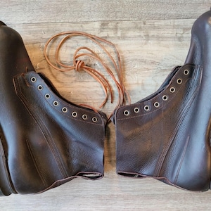 May include: A pair of brown leather work boots with multiple eyelets for laces. The boots have a rugged sole and are displayed with their laces. The boots are made of a dark brown leather.