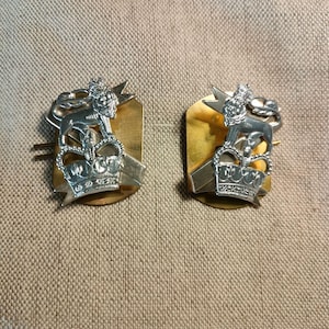 May include: Two gold-coloured metal badges with a silver lion, crown, and star design. The badges are on a beige fabric background.