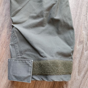 May include: Close-up of a section of olive green fabric, possibly a jacket or pants. The fabric has a matte finish and is slightly creased. A rectangular patch and a strip of hook-and-loop fastener are visible.