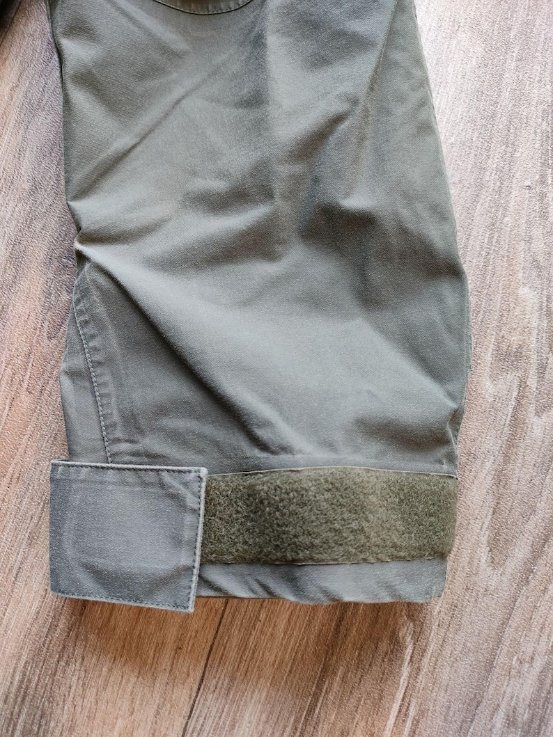 May include: Close-up of a section of olive green fabric, possibly a jacket or pants. The fabric has a matte finish and is slightly creased. A rectangular patch and a strip of hook-and-loop fastener are visible.