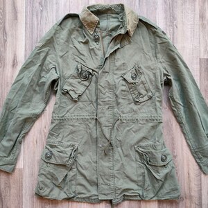 Op de afbeelding: Een olijfgroene jas in militaire stijl met meerdere zakken en een knoopsluiting. De jas heeft een kraag en lange mouwen. De stof lijkt een duurzame, mogelijk katoenen mix te zijn. De jas ligt plat op een houten oppervlak.