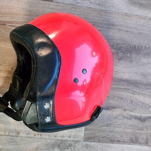 May include: A bright red motorcycle helmet with a black leather trim and chin strap. The helmet has a rounded shape and is secured with a black strap and buckle. The helmet is on a wooden surface.
