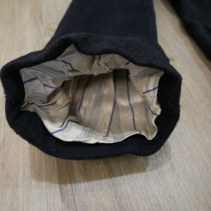 May include: Close-up of the inside of a black wool coat sleeve. The sleeve is lined with a beige fabric with thin blue stripes.